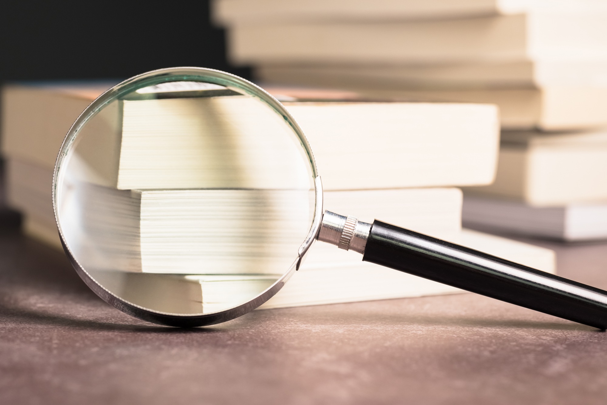 A magnifying glass rests on a table, focusing on stacked books. This image symbolizes exploration and detailed examination of literature.