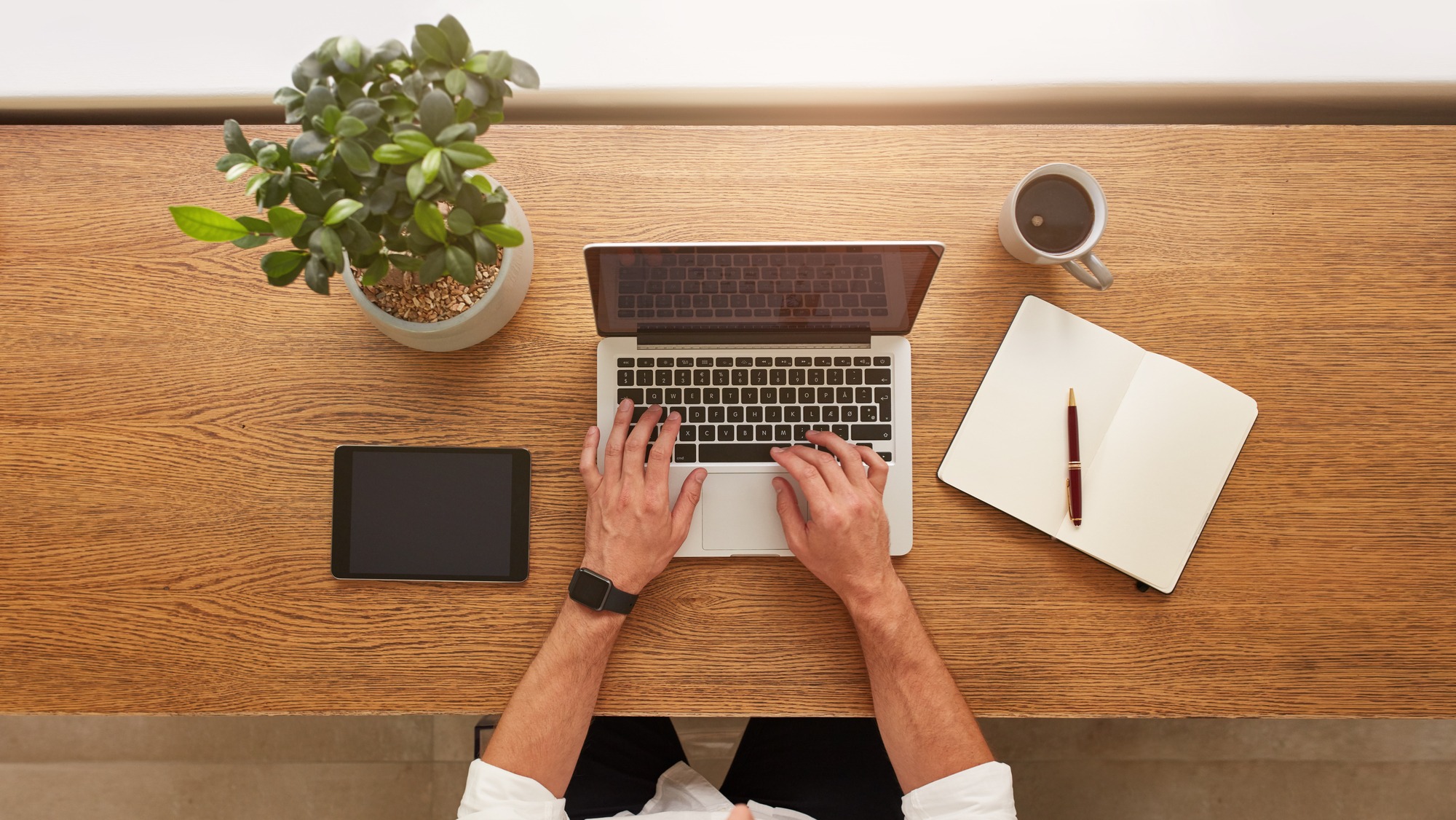 Person typing on a laptop at a wooden desk, accompanied by a tablet, notebook, pen, coffee mug, and a small potted plant.