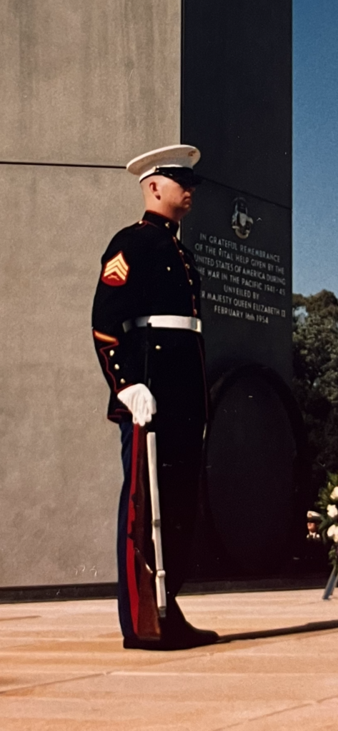 Uniformed Marine standing at attention next to a memorial dedicated to American aid in the Pacific during WWII, with flowers nearby.