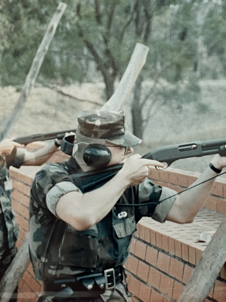 Soldier in camouflage prepares to shoot over a brick barrier, wearing headphones and aiming a shotgun, amid a wooded training environment.