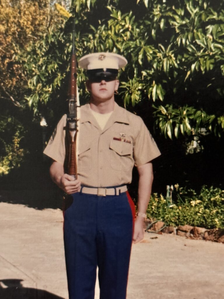 A Marine in dress uniform stands at attention, holding a rifle, against a backdrop of lush greenery. The image showcases military discipline and pride.
