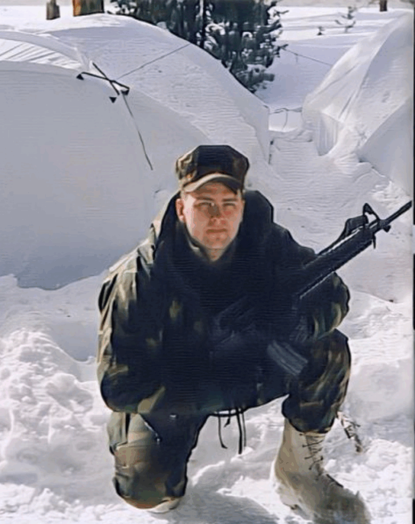 Soldier in winter camouflage gear crouches in snow, holding a rifle, with tents visible in the background. Demonstrates military readiness in a snowy environment.