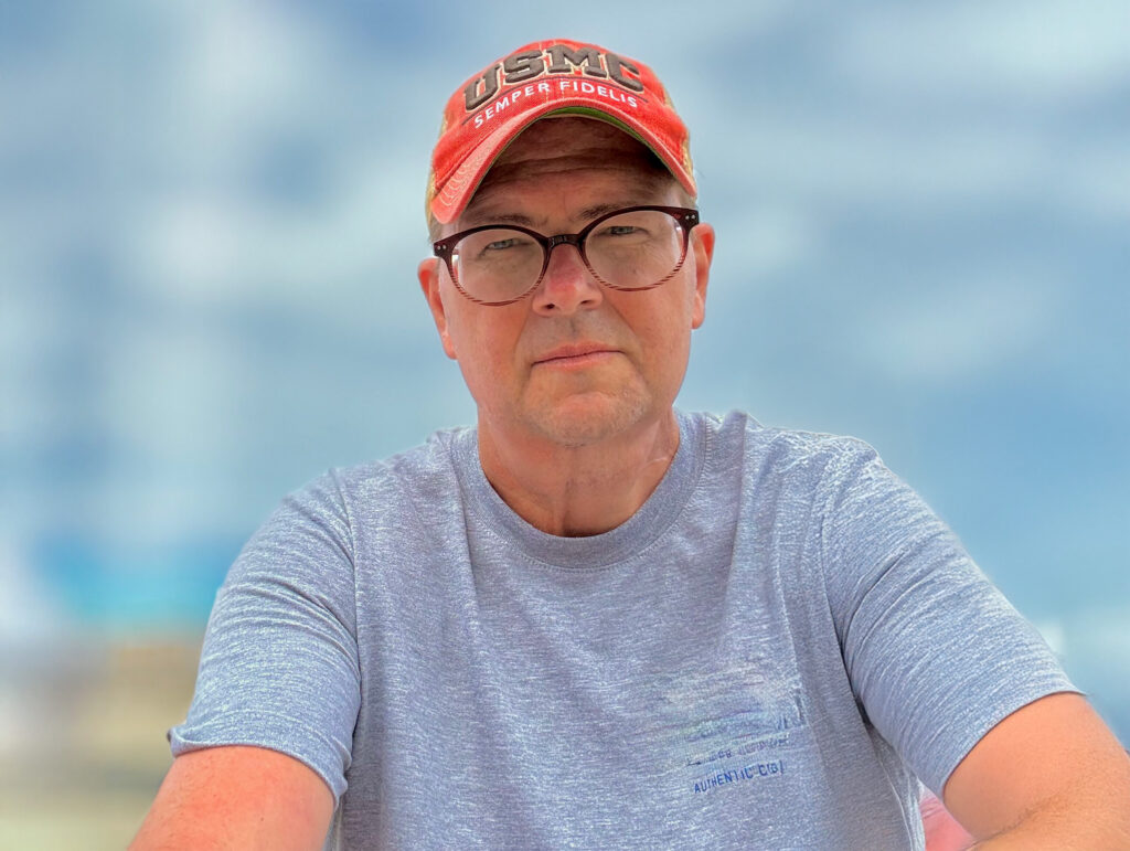 Man wearing a red USMC hat and gray t-shirt, looking directly at the camera against a blurred sky backdrop.