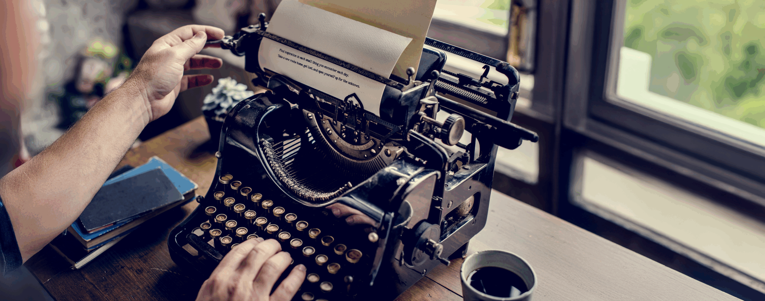 A hand operates a vintage typewriter, with a partially typed sheet emerging. Nearby, stacked notebooks and a coffee cup enhance the cozy writing atmosphere.